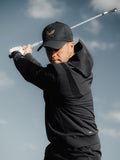 Golfer in black ProTek hoodie mid swing, wearing a cap against a clear sky