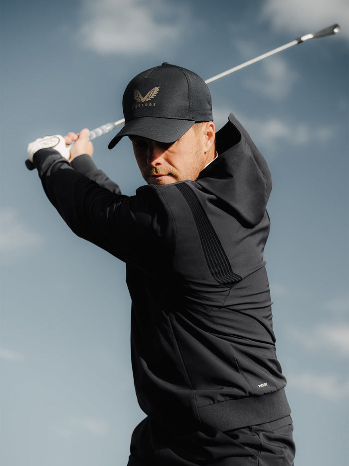 Golfer in black ProTek hoodie mid swing, wearing a cap against a clear sky