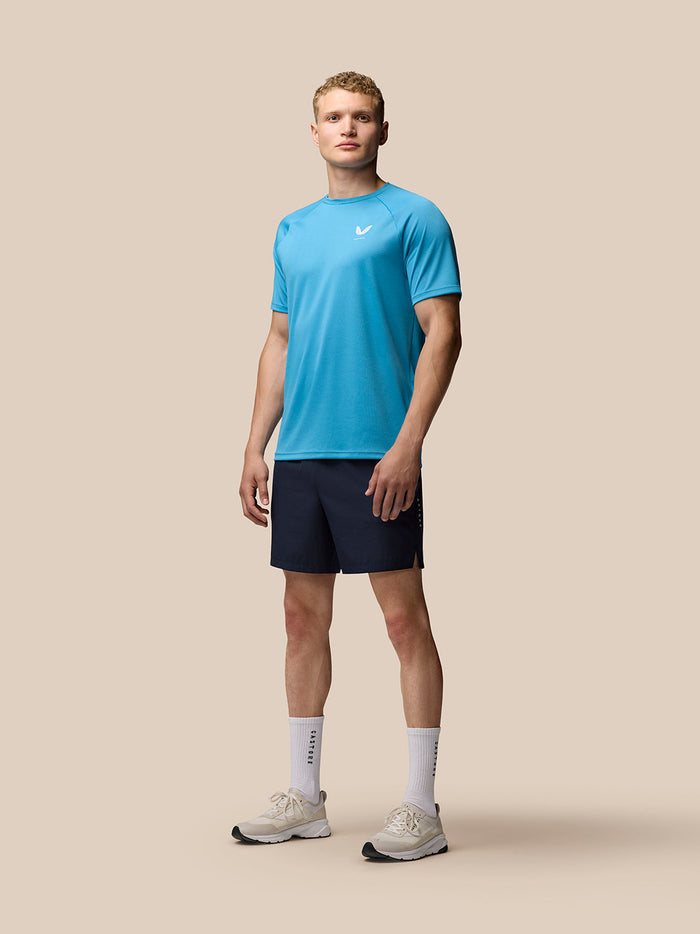 Full-body shot of a male model wearing a turquoise athletic T shirt and navy 7 inch shorts with side branding and sneakers.