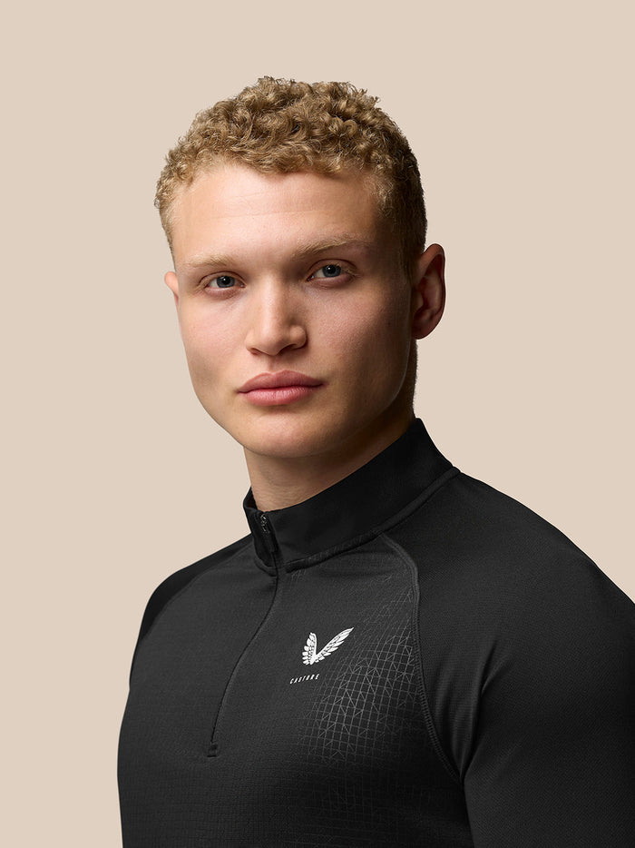 Three-quarter rear view of male model in black Adapt Print Quarter Zip Top showing subtle pattern and small white Castore logo.