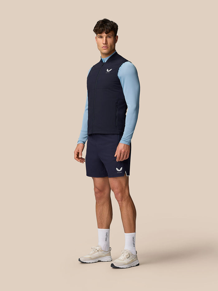 Model wearing navy Apex Training Gilet over a blue long sleeve top and navy shorts posing against a neutral backdrop
