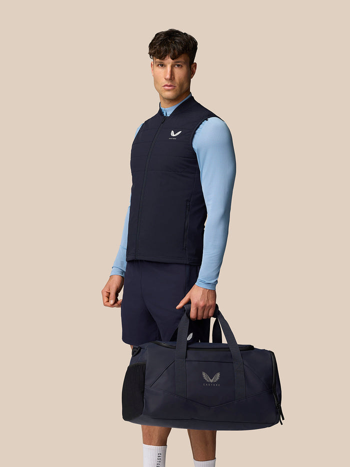 Full body view of a man in navy Apex Training Gilet over a light blue top with navy shorts and a bag