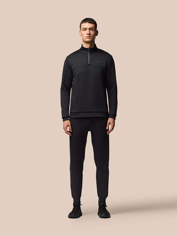 Full body shot of a man wearing a black Castore Flex Lifestyle quarter zip top with matching pants against a neutral beige background
