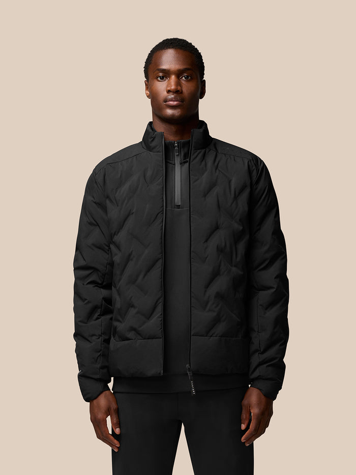 Male model wearing a black padded jacket with stand collar and zip front, featuring side pockets and left shoulder branding