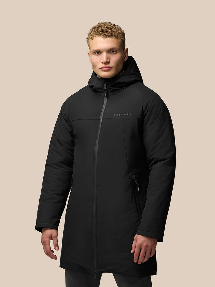 Male model wearing black longline wadded jacket with hood and full front zipper against a beige backdrop
