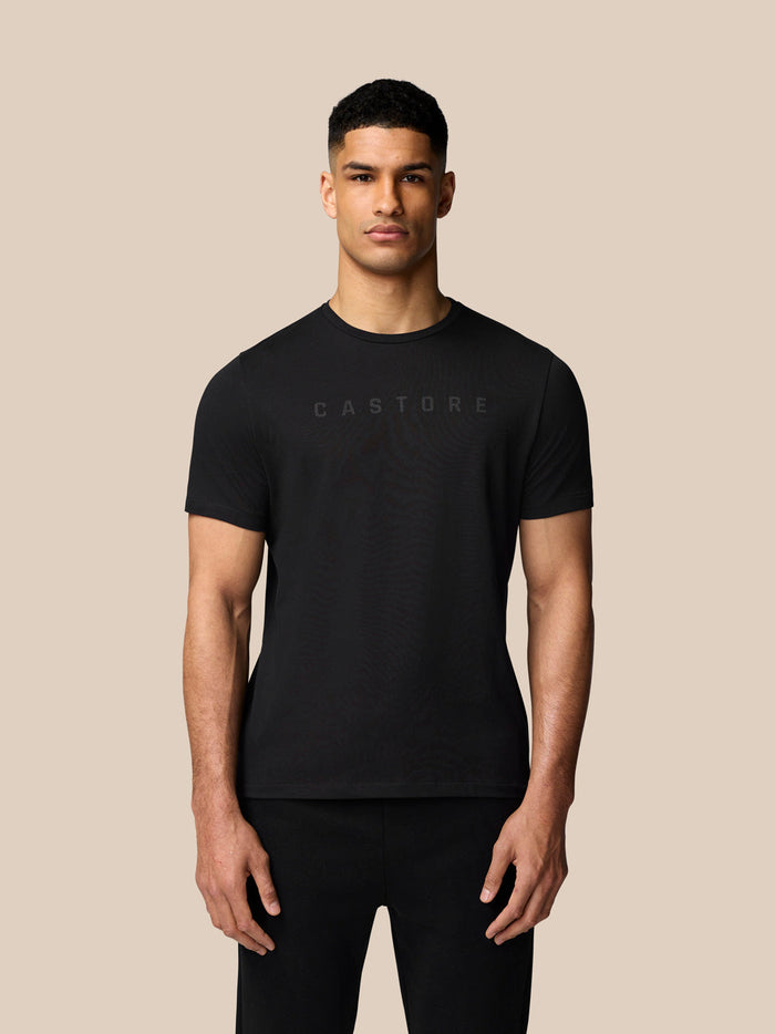 Male model wearing black short sleeve graphic T shirt with a subtle Castore logo on the chest against a beige background.