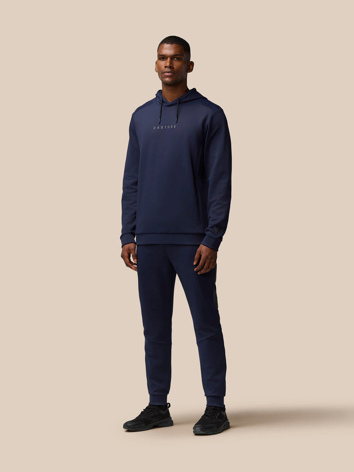 Male model wearing navy Castore Flex Lifestyle Thermo Pant tracksuit with hoodie, full-body view.
