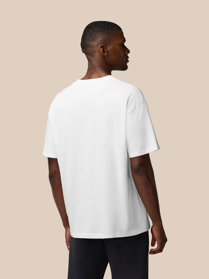 Side view of a male model wearing a white T shirt with round neck and black joggers on a beige backdrop