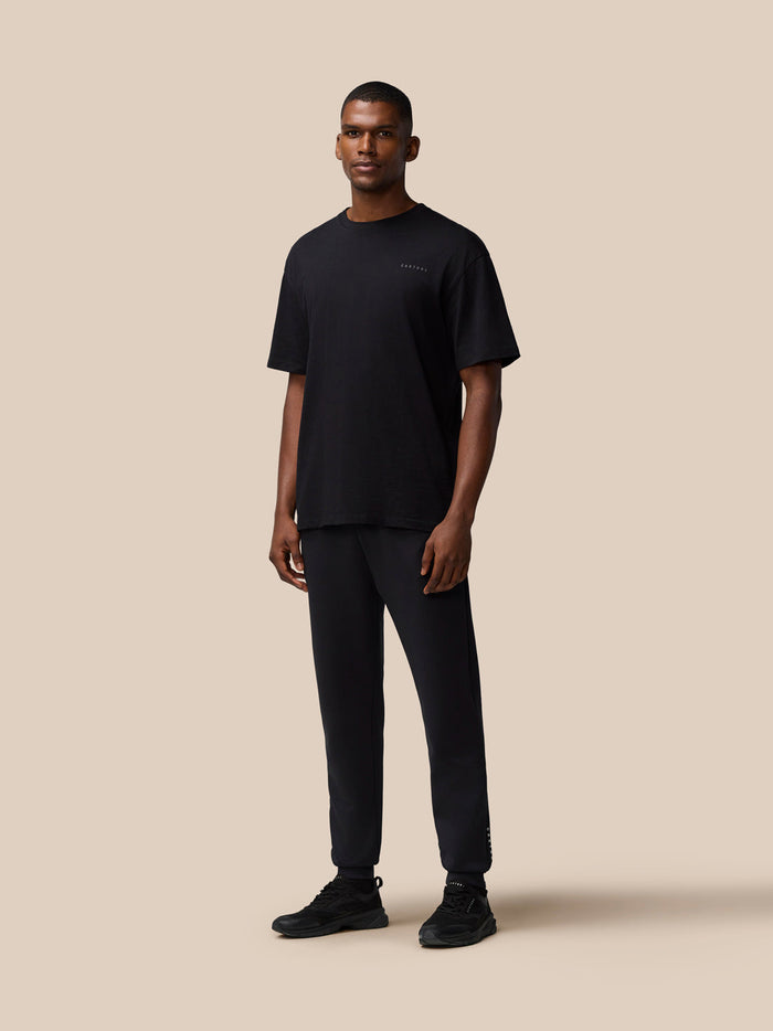 Full body shot of a male model in a black Flex Cotton T Shirt and matching pants, standing against a tan background.