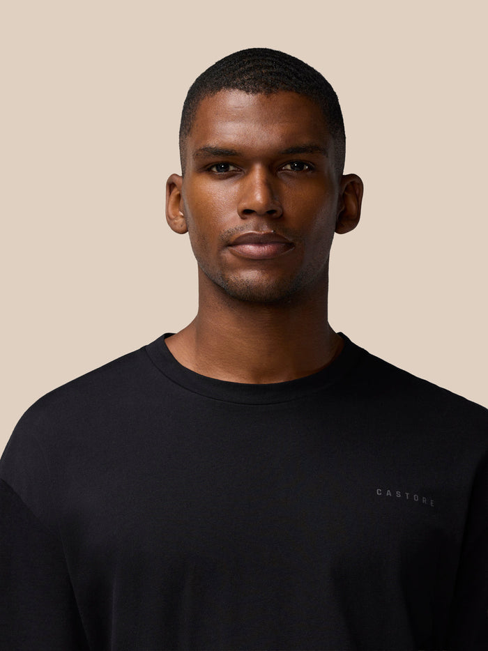 Close up of a male model wearing a black Flex Cotton T Shirt with a round neckline against a tan background.