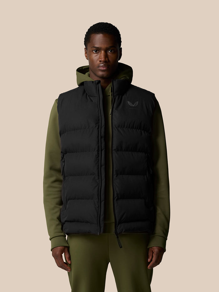 Male model wearing black heavyweight padded gilet over olive green hoodie against a beige backdrop