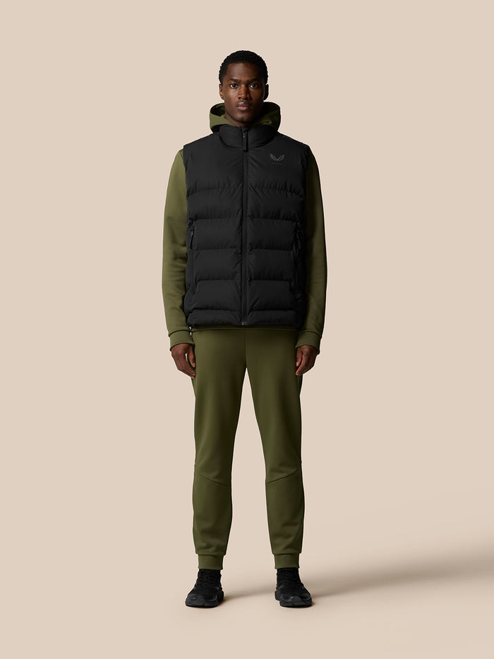 Full body shot of a model wearing a black heavyweight padded gilet over an olive green hoodie and joggers.