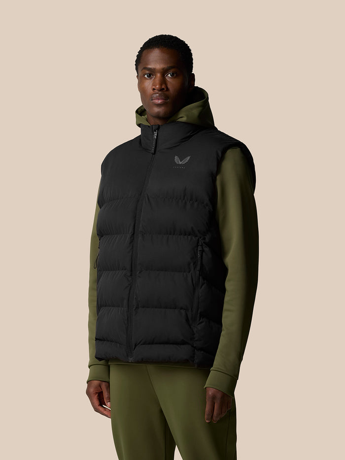 Mid shot of a model in the black padded gilet over an olive green hoodie, front view with zip closure.