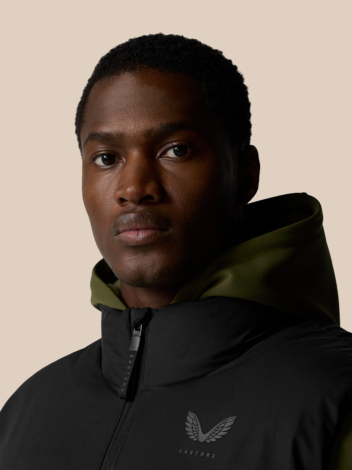 Close-up of a model wearing the black padded gilet with hood, chest logo visible against a beige background.