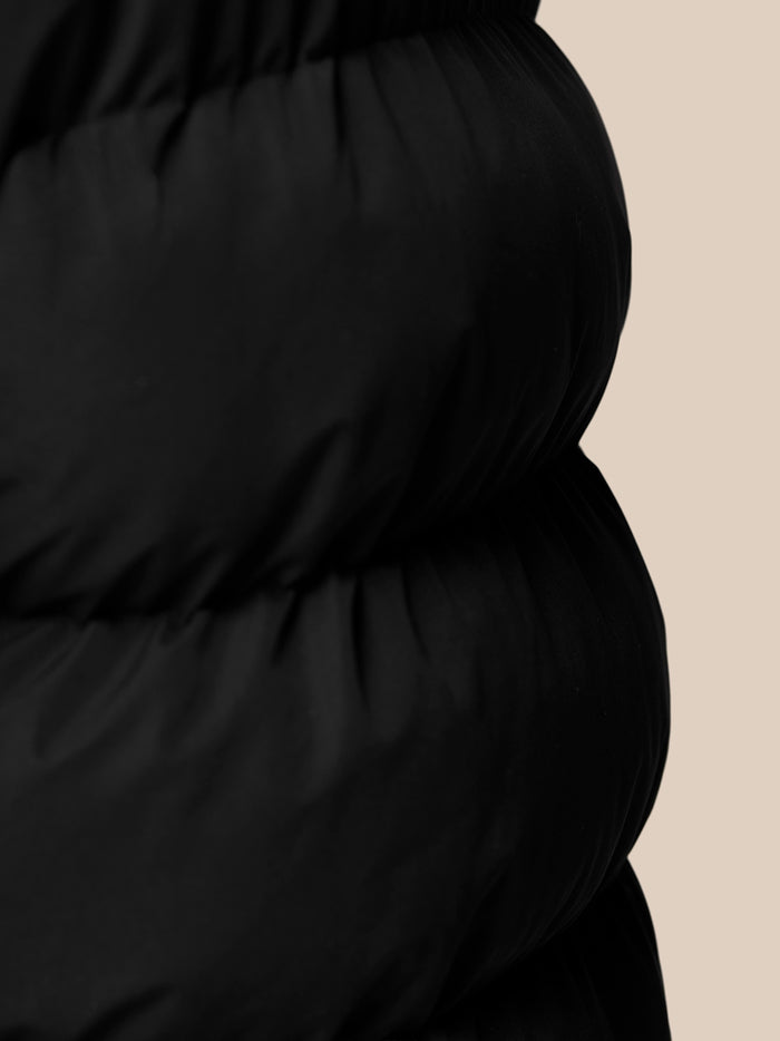 Close-up of black padded gilet with gathered waist against a beige backdrop.