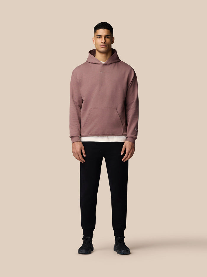 Full body shot of a female model wearing mauve Adapt Lifestyle Hoodie with front pocket against a beige background.