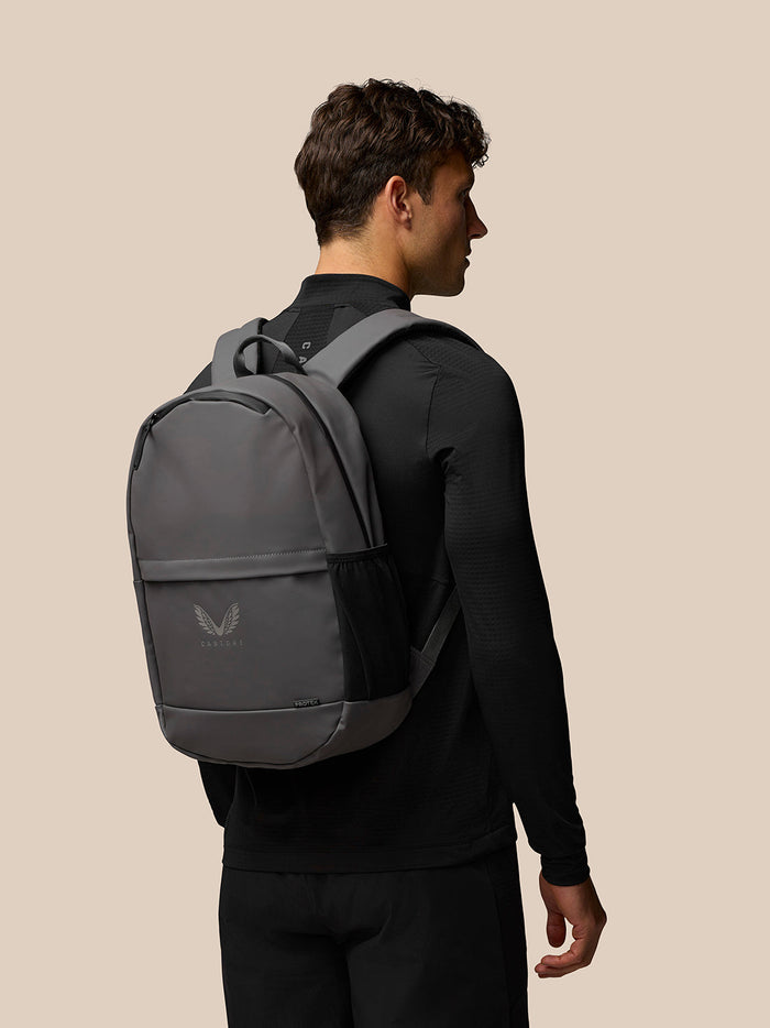 Charcoal grey Castore core backpack worn by a model showing padded back and reflective logo.