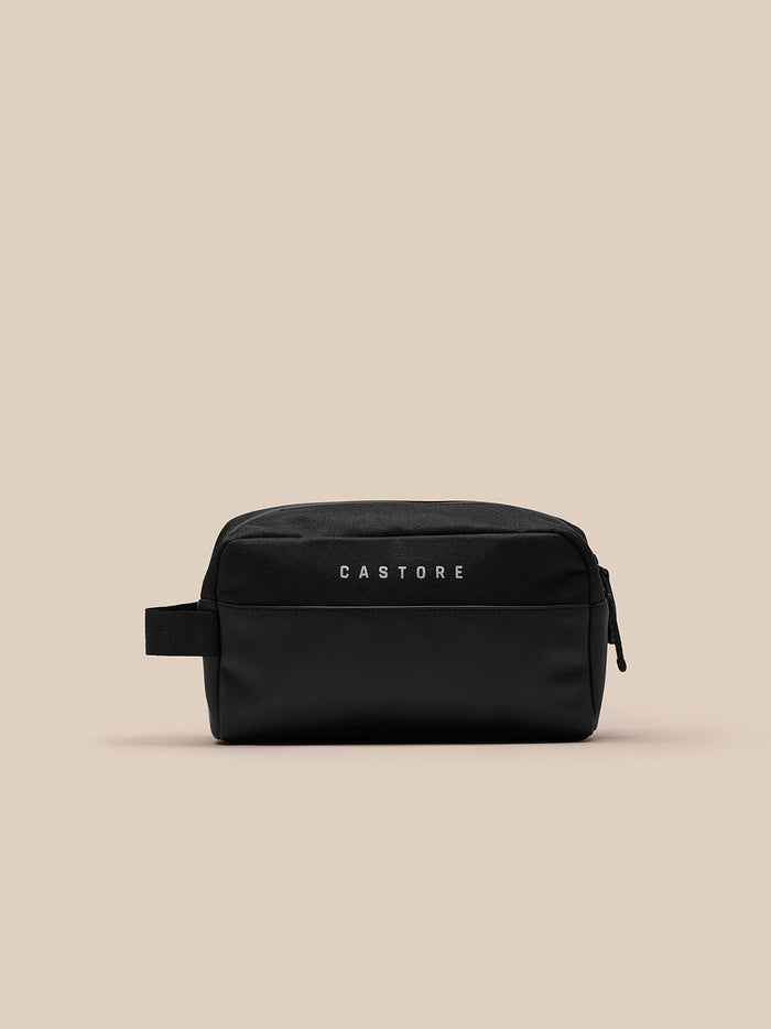 Black Active Wash Bag by Castore with front pocket and side strap against a beige background.