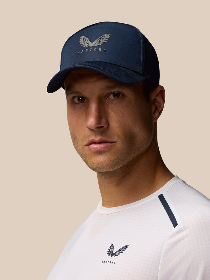 Front-on view of a navy Castore cap with wing logo worn by a model in a white shirt