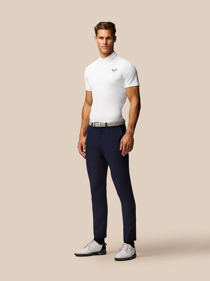 Male model wearing white Golf Club Essential Polo, navy trousers and white golf shoes with Castore logo on the chest.
