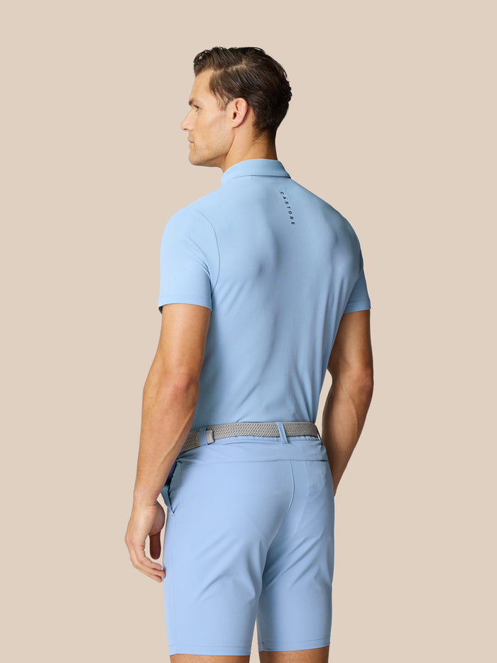 Back view of a man in a sky blue golf polo and matching shorts with a light belt; Castore branding runs along the upper back.