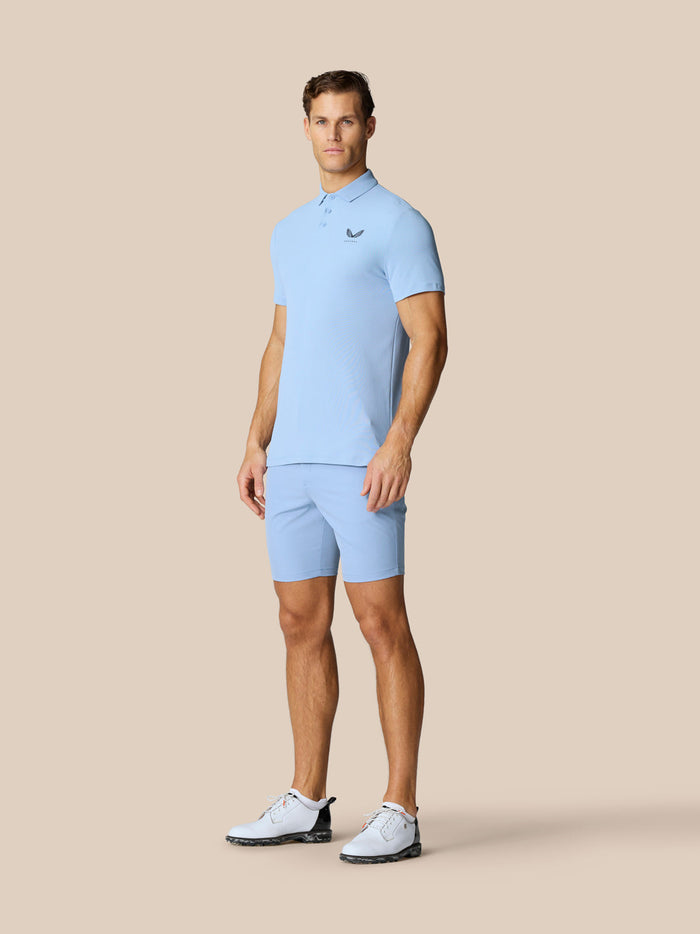 Male model in sky blue Golf Club Essential Polo and matching shorts with golf shoes posed against beige background