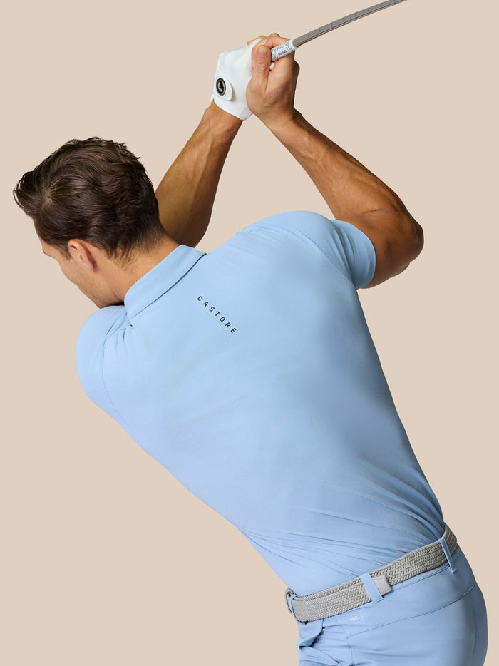 Side angled shot of a man in sky blue golf polo and shorts swinging a golf club with Castore logo on the back of the polo.