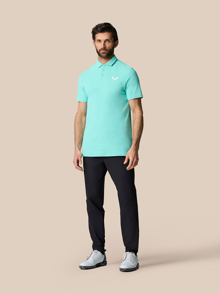 Full-body view of a man wearing light aqua Golf Club Essential Polo with dark trousers and gray golf shoes with orange accents.