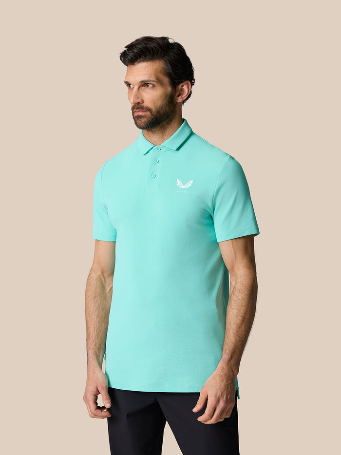 Full-length shot of a man in light aqua Golf Club Essential Polo with Castore logo on the chest against a beige backdrop.