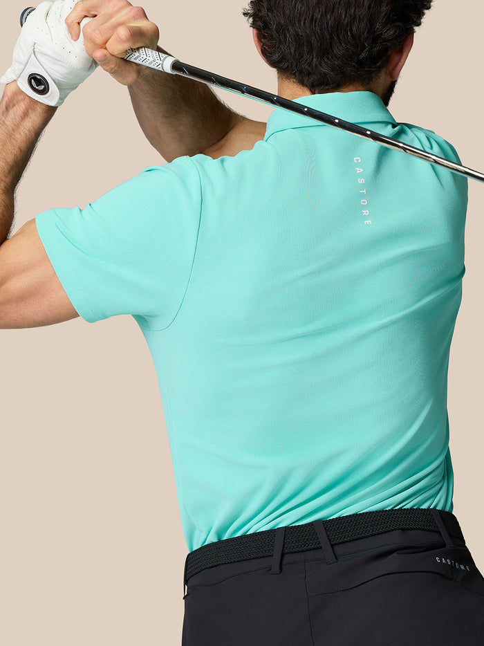 Athlete in a light aqua Golf Club Essential Polo mid swing with a golf club and Castore branding on the back.