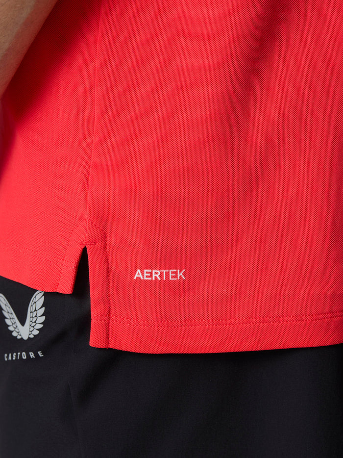 Full-body view of a man in a red Golf Club Essential Polo with a left chest Castore logo, paired with dark trousers.