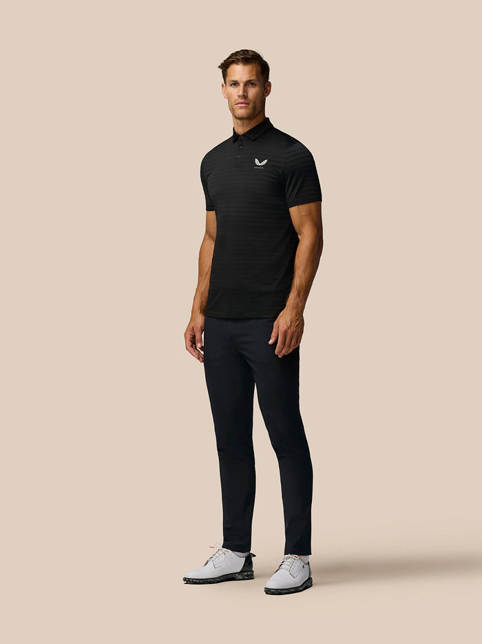Full-length shot of a man in a black Pinnacle Pique Stripe Polo with dark trousers and white golf shoes