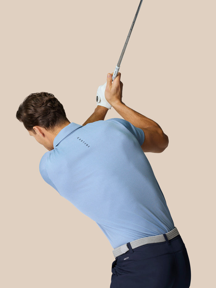 Male model wearing sky blue Engineered Knit Polo viewed from behind during a swing with branding at the upper back.