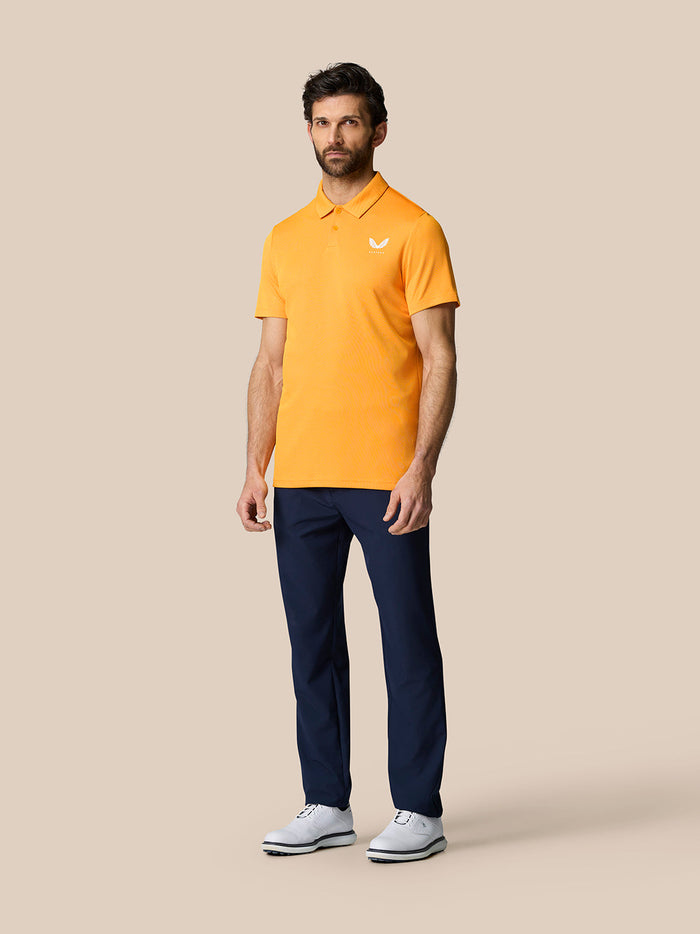 Full-body shot of a model in mango orange polo with navy pants and white shoes against a beige backdrop.