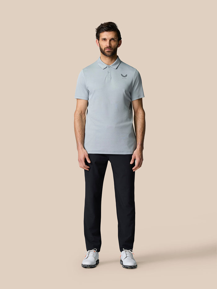Full-body shot of a man in a light blue polo shirt, navy pants, and white sneakers against a beige background.