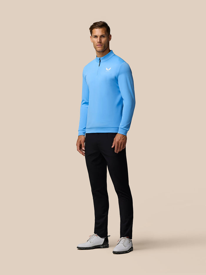 Full body view of a man wearing a light blue Golf Club Classic 1/4 Zip Top with black trousers and white sneakers in a three-quarter pose
