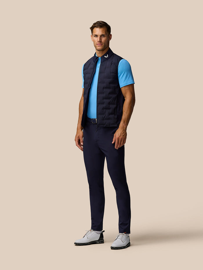 Male model in navy Pinnacle gilet over a light blue polo, angled side view showing front zip and stand collar