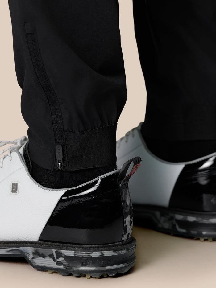 Close up of black golf trousers at the ankle showing a zipper closure and elastic cuff beside a white sneaker.