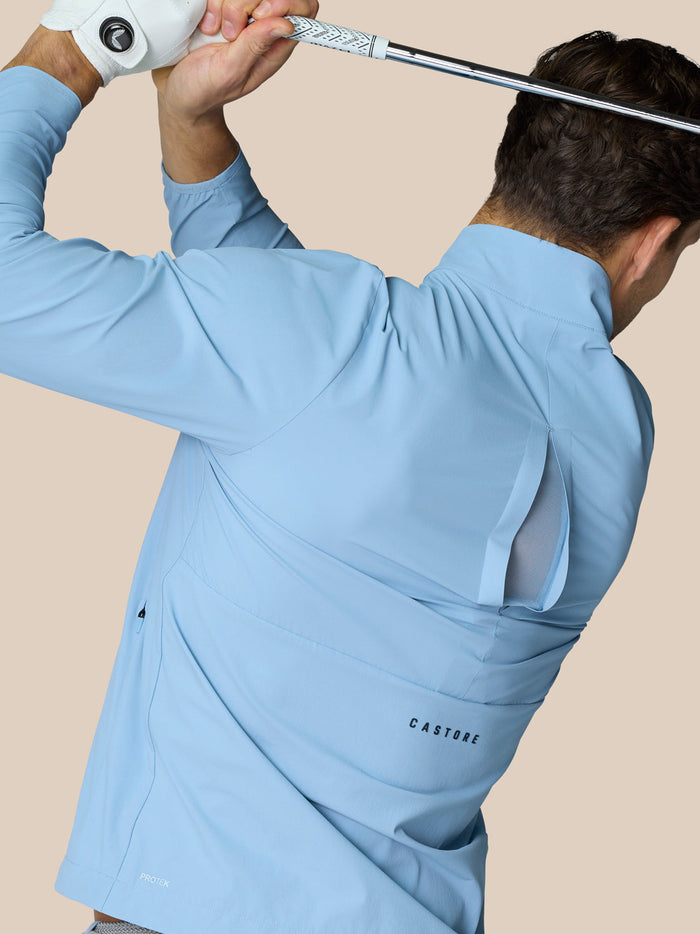 Back view of a man wearing a sky blue Castore Golf Flex Jacket while swinging a golf club.