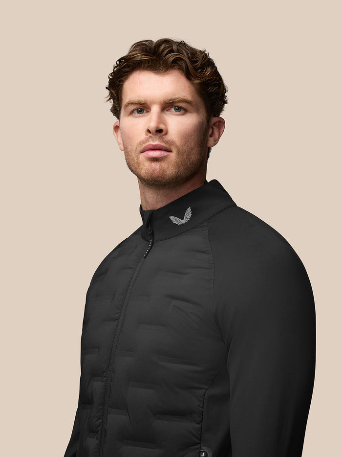 Full body shot of a man wearing the black Golf Pinnacle Jacket with stand collar and front zip against a beige backdrop