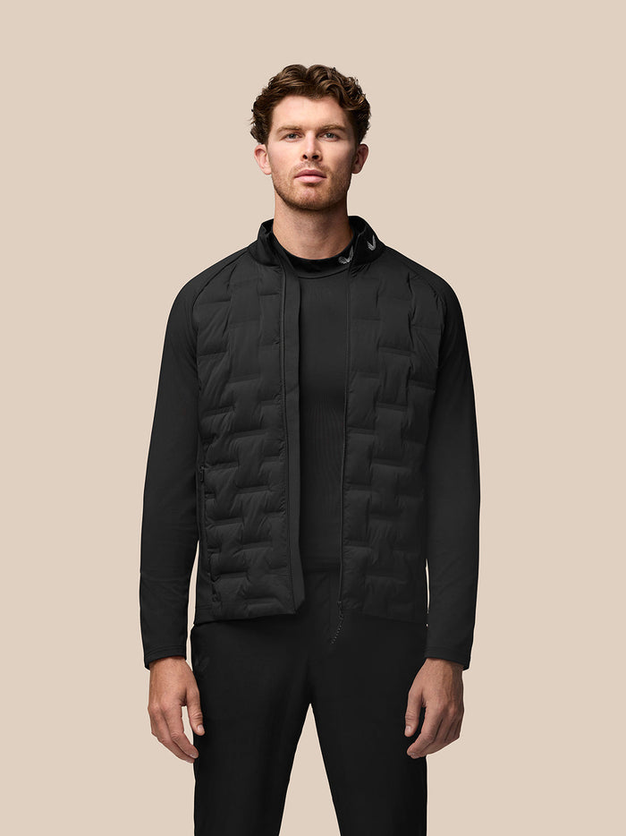 Model wearing black Golf Pinnacle jacket with quilted front panels, stand collar and full zip against a beige backdrop.