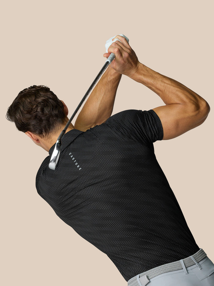 Back view of a golfer in a black dotted polo mid swing with a golf club and Castore branding on the back