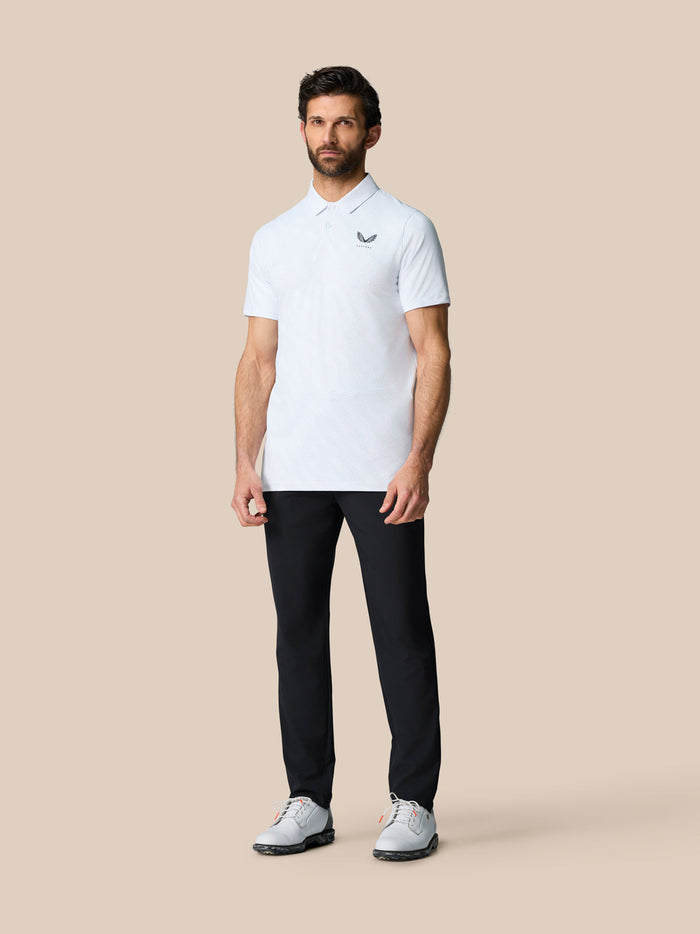 Full body of a man in a white polo and dark trousers with Castore logo on the left chest, posing against a beige backdrop.