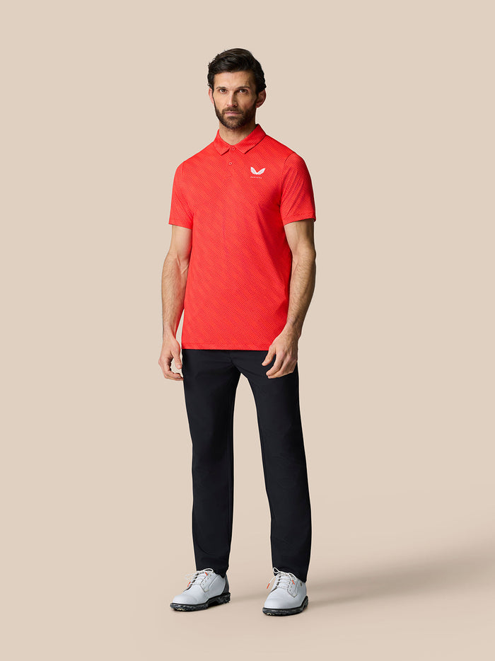 Full-length shot of a man in a red Club Graphic Polo with a white Castore logo on the chest, black trousers and white sneakers.