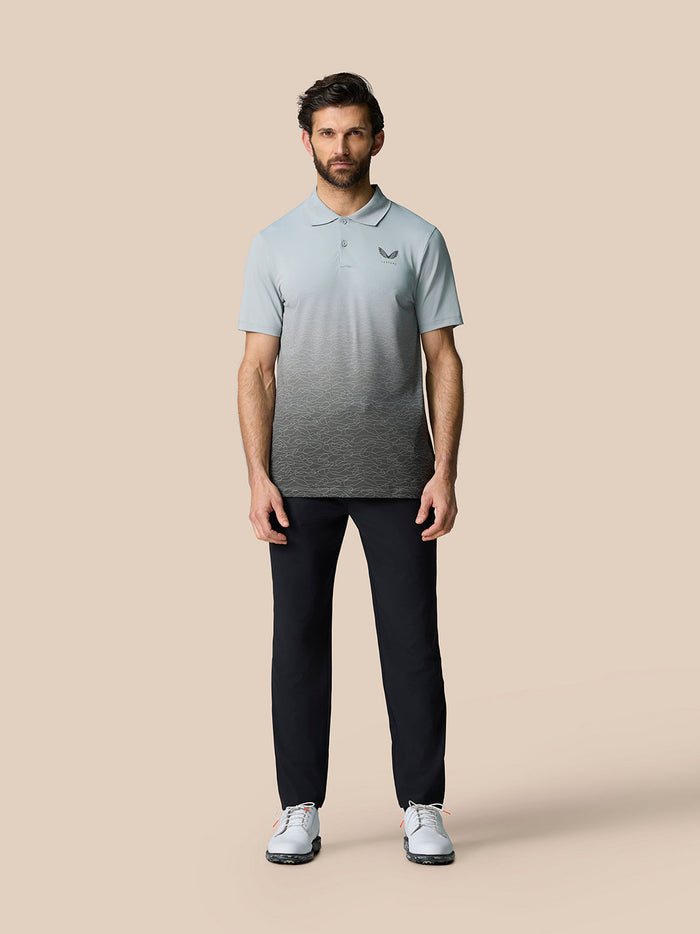 Full body shot of a man in gradient Club Graphic Polo from light blue to charcoal, Castore logo on chest, navy trousers and white sneakers.