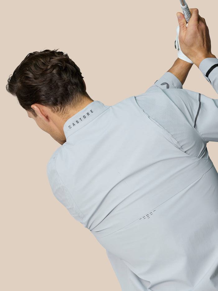 Back view of a man wearing a light grey Castore Golf Pinnacle LVS Woven Jacket with venting panels and Castore logo on the collar
