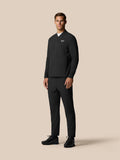 Full-body view of a male model in a black Castore golf jacket with matching trousers against a beige backdrop.