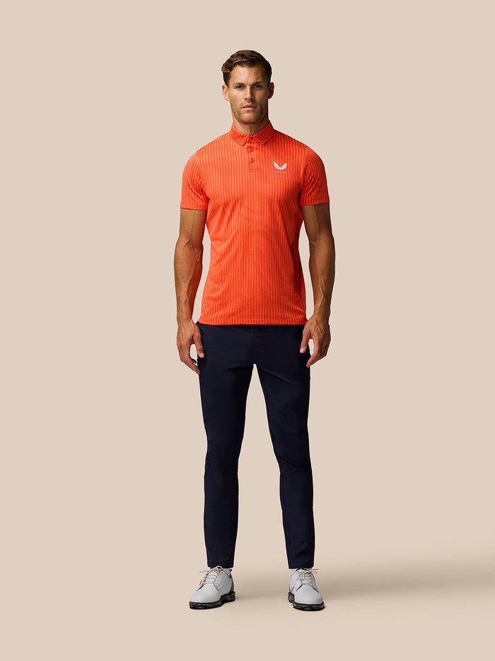 Male model wearing bright orange striped golf polo with Castore logo, navy pants and white sneakers.