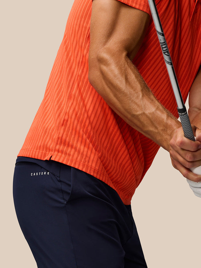 Side view of a man in a bright orange striped Golf Pinnacle Glow Polo holding a golf club.
