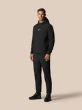 Male model in a black golf performance hoodie and matching pants with a small Castore logo on the chest against a beige studio backdrop